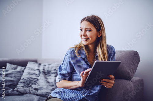 Fotografía Young charming smiling caucasian brunette in stripped shirt sitting on sofa in living room, holding tablet and looking away