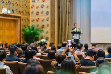 Back side of audience taking photo by cellphone the Speaker with podium and presentation via projector on the stage in the conference hall, business and education concept