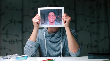 Fototapete - A guy sitting at a table holds tablet screen in his hands in place of his head.