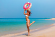 Tourist vacation woman jumping and joying with funny pink donut in bikini and hat. Beach model laughing in Dubai. Travel woman in famous destination UAE. Nice sand beach clear water in Jumeirah Dubai.