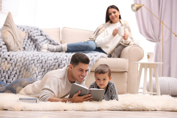 Wall Mural - Father and his son reading books while mother resting on sofa at home. Winter vacation