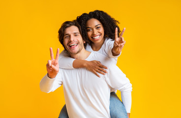 Poster - Joyful interracial couple embracing and showing peace sign over yellow background