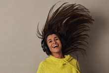 Amazed Youthful Attractive Female Brunette In Yellow Sweater And Headphones Looking At Camera And Jumping While Listening Music On Gray Background In Studio