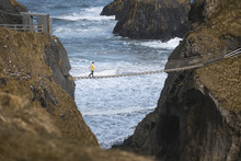 From Above Side View Of Traveler Passing Over Carric A Rede Rope Bridge Suspended Between Rocky Cliffs And Sea Waves Crashing On Rocks In Background