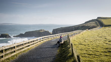 Woman Sitting Resting During A Walk On Narrow Rural Road Along Green Field On Sunny Spring Day At Coastline In Northern Ireland