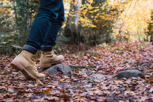 Crop Legs In Jeans And Brown Boots Of Hiker On Rocky Spangled Of Golden Fallen Leaves Path With Autumn Forest On Background