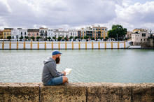 Side View Of Bearded Male Art Student In Casual Clothing Sitting With Crossed Legs On Rocked Fence Of Quay And Drawing Sketches In Small Album With River And Bridge On Background