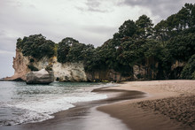Landscape Of Sandy Beach And Ocean Waves Beside Tropical Cliffs Overgrown With Greenery On Cloudy Day At Coromandel Peninsula In New Zealand