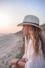 Side View Of Graceful Woman With Long Curly Hair In Hat Leaning On Fence And Looking Away At Seaside