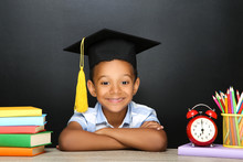 Young African American School Boy Sitting At Desk With Books, Pencils And Alarm Clock On Black Background