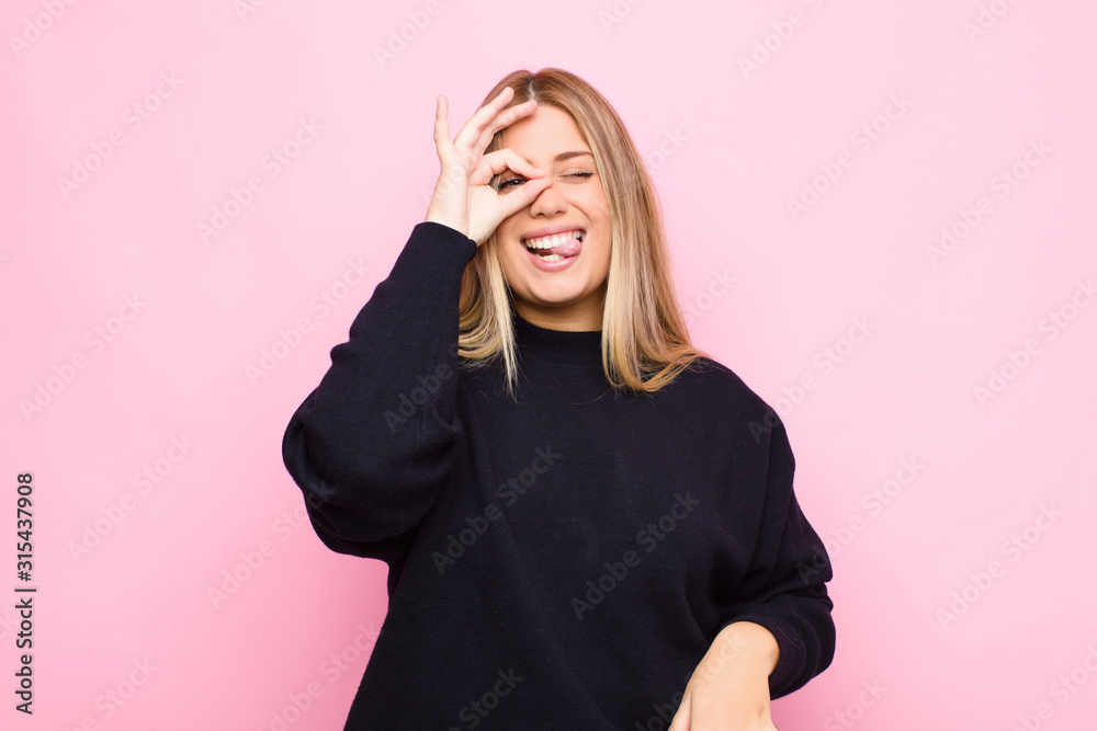 young blonde woman smiling happily with funny face, joking and looking through peephole, spying on secrets against flat wall - obrazy, fototapety, plakaty 