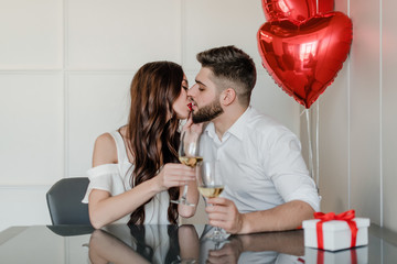 Wall Mural - happy handsome man and woman drink white wine at home with present in gift box and red heart shaped balloons at home