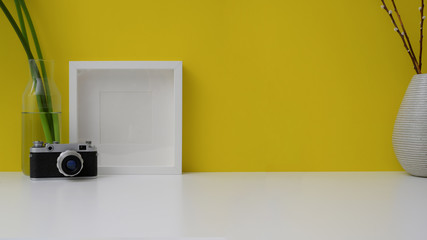 Cropped shot of workspace with copy space, mock up frame, camera and vase on white table with yellow wall background