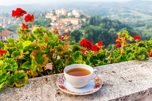 One Black Tea Cup On Balcony Terrace Railing By Red Geranium Flowers Outside In Italy With Mountain Of Chiusi Cityscape In Tuscany View