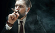 © MYDAYcontent - Bearded businessman inhaling cigar smoke wearing dark suit, tie, and watch against black background
