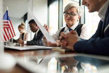 Wall Mural - Smiling senior businesswoman in eyeglasses sitting at the table reading report and talking to her colleague at conference