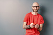 Handsome hairless Caucasian smiling man with beard, glasses, red T-shirt holding phone in hands on gray studio background
