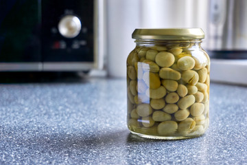 Single glass jar with canned broad beans on kitchen top with microwave. Image with copy space and selective focus.