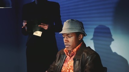 Wall Mural - Close-up of accused black man in hat covering his face with his hands during interrogation. In the background there is a female detective writing notes.