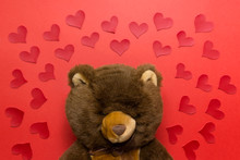 Funny Valentine Day Greeting Card With Brown Plush Teddy Bear And Many Fabric Red Hearts On Red Background.