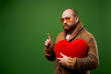 Man In Pink Glasses Holds Toy Heart While Standing On Empty Green Background In Studio