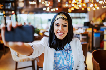 Charming modern smiling muslim woman with scarf on head sitting in cafe and taking selfie for social media. Millennial generation.