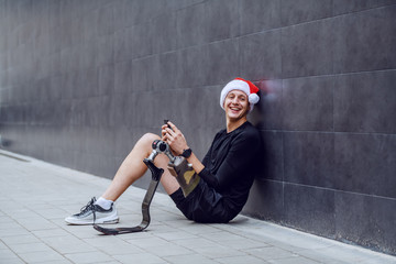 Wall Mural - Cheerful Caucasian sportsman with artificial leg and with santa hat on head sitting on ground, leaning on wall and sending wishes for christmas over his smart phone.
