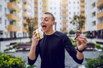 Wall Mural - Attractive sportsman standing outdoors and choosing between candy and fresh banana. In background are buildings.