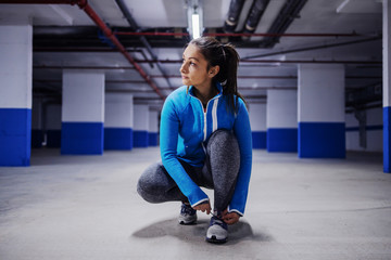 Young fit attractive caucasian sportswoman in tracksuit crouching and tying shoelace. Urban life concept.