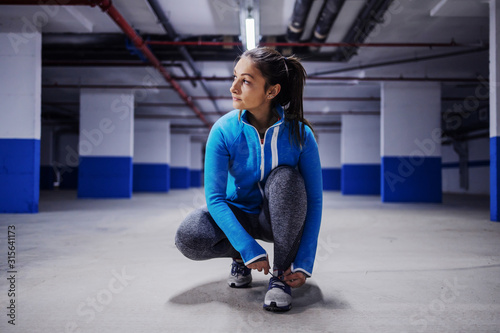 Young fit attractive caucasian sportswoman in tracksuit crouching and tying shoelace Tableau sur Toile