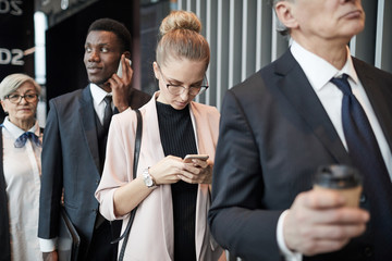 Wall Mural - Young businesswoman in eyeglasses typing a message on her mobile phone while standing in the queue with other people at the airport