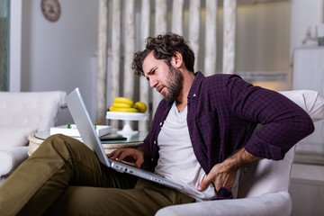 Tired man freelance worker stretch in sofa suffer from sitting long in incorrect posture, male employee have back pain or spinal spasm working in uncomfortable position. Sedentary life concept