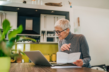 Wall Mural - Smiling senior woman working with documents and laptop in the kitchen at modern home.