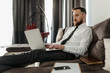 © Marat - A bearded male freelancer in a shirt and tie is comfortably spread out on the couch, sitting in his living room, and working on a laptop