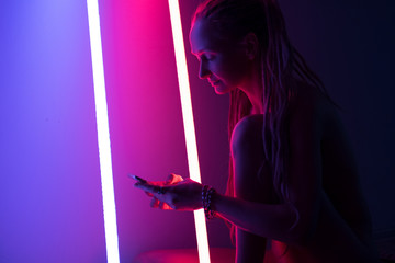 Studio portrait of woman seating in red and purple coloured lamps light using cell phone