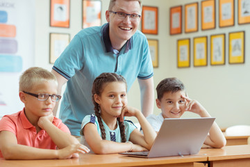 Wall Mural - Young handsome teacher with group of clever children working with laptop during a lesson