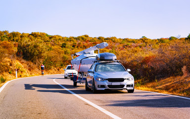 Canvas Print - Car trailer with yacht or motorboat on road at in Sardinia Island, Italy summer. Minivan with motor boat on motorway on holidays at highway. Olbia province. Mixed media.