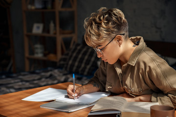 Education. Young woman short hair in glasses sitting at desk studying reading book taking notes concentrated close-up