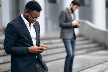 Wall Mural - Young handsome african american businessman is walking on stairs and typing something on his phone with smile, outdoors