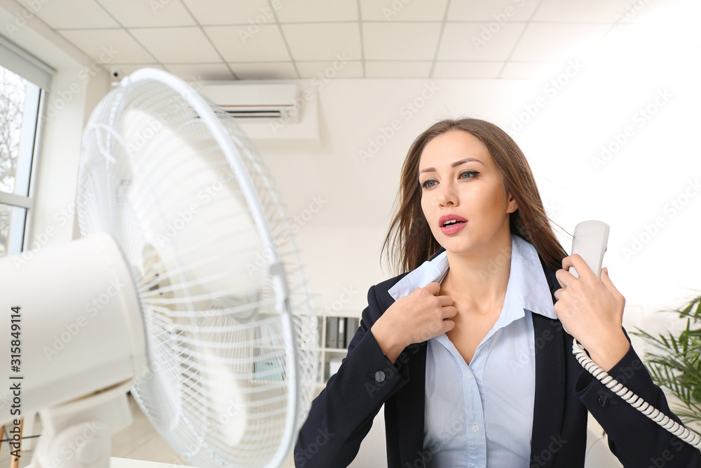 Young woman using electric fan during heatwave in office