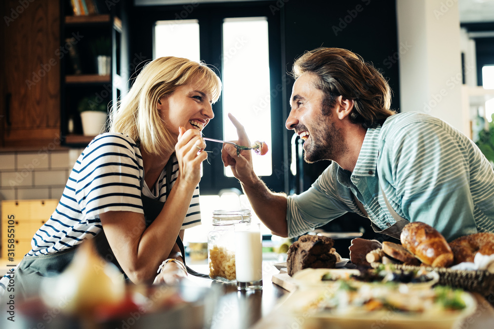 Beautiful young couple is smiling while cooking together in kitchen at home - obrazy, fototapety, plakaty 