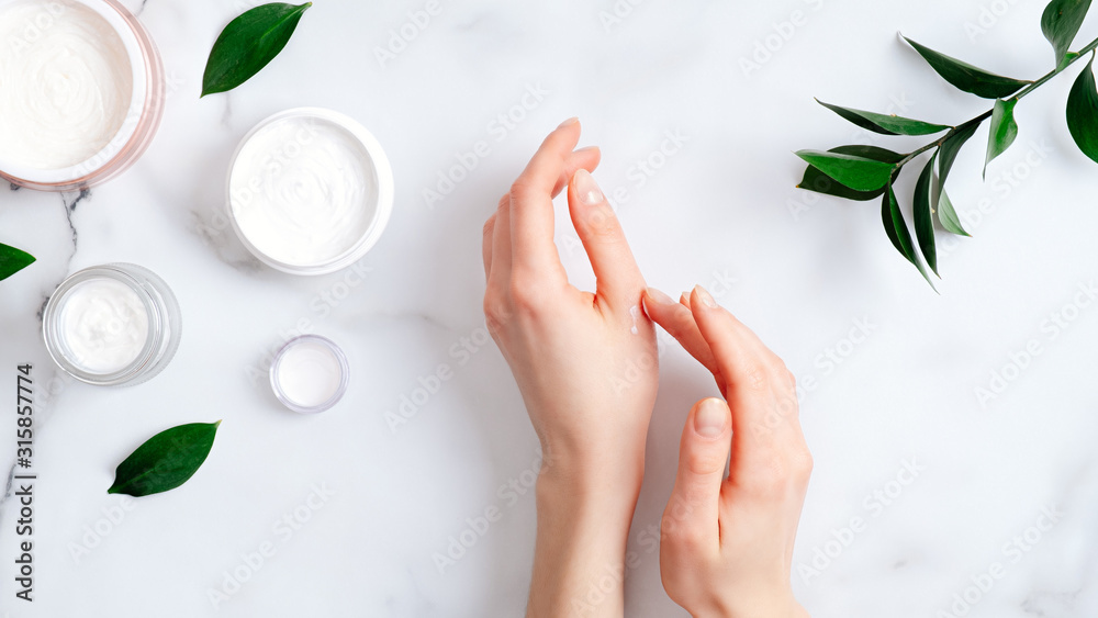 Cosmetic cream on female hands, jars with milk swirl cream and green leaves on white marble table. Flat lay, top view. Woman applying organic moisturizing hand cream. Hand skin care concept - obrazy, fototapety, plakaty 