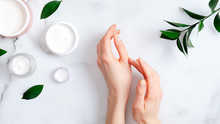 Cosmetic Cream On Female Hands, Jars With Milk Swirl Cream And Green Leaves On White Marble Table. Flat Lay, Top View. Woman Applying Organic Moisturizing Hand Cream. Hand Skin Care Concept