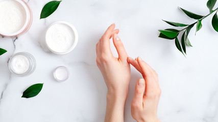 Wall Mural - cosmetic cream on female hands, jars with milk swirl cream and green leaves on white marble table. f
