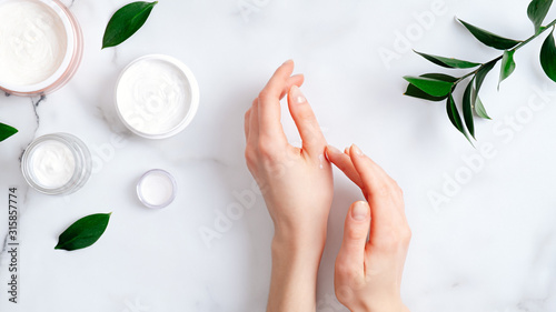 Fotografía Cosmetic cream on female hands, jars with milk swirl cream and green leaves on white marble table