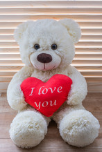 Cute Teddy Beige Bear Sits On A Background Of Wooden Blinds. He Holds A Big Red Heart With The Text I Love You