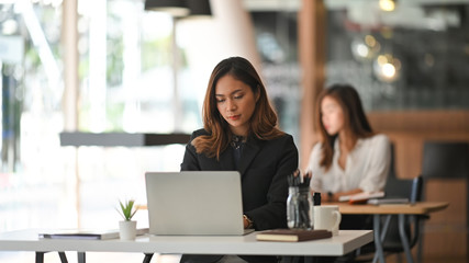 Wall Mural - Photo of young business woman in look good black suit typing on her white laptop while sitting at work desk in the modern office. Sitting over colleague background.