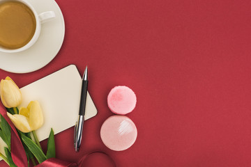 Wall Mural - top view of tulips with silk ribbon, macarons and empty notebook near coffee isolated on red