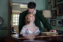 Vintage Couple In Period Costume Sitting Around Piano Playing Tunes With Man Kissing Woman In The Head