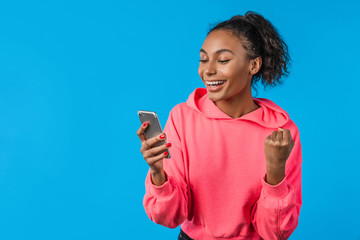 Excited african woman celebrating with mobile phone isolated over blue background